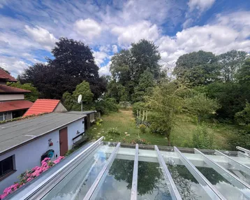 Blick von der Glasüberdachung auf den weitläufigen Garten mit altem Baumbestand und viel Grün.