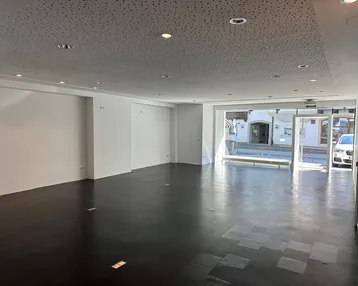 Geräumiger, leerer Geschäftsraum mit dunklem Boden, weißen Wänden und großer Schaufensterfront zur Straße.