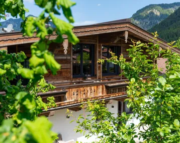 Traditionelles Holzhaus im alpinen Stil, umgeben von üppigem Grün und Bergen im Hintergrund.