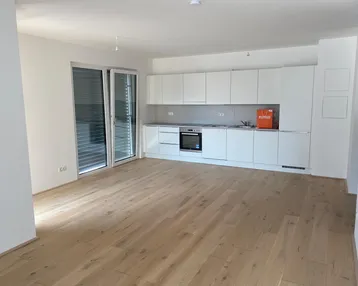Heller offener Wohnbereich mit integrierter weißer Küche, Parkettboden und Zugang zum Balkon.