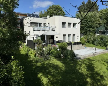Modernes Wohnhaus mit großem Garten, Terrasse und üppiger Vegetation unter blauem Himmel.