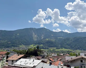 Panoramablick über eine Stadt mit roten Dächern und grünen Bergen unter blauem Himmel.