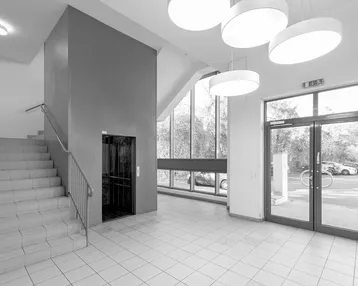 Heller Eingangsbereich mit Treppe, Aufzug und großen Fenstern, die viel Tageslicht hereinlassen.