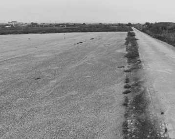 Weitläufige, flache Fläche, möglicherweise ein ehemaliger Flugplatz oder Parkplatz, mit spärlicher Vegetation.