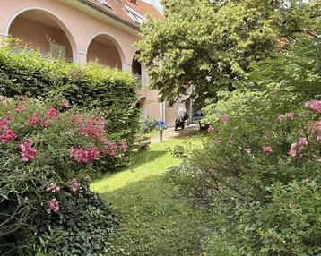 Grüner Gartenbereich mit blühenden Sträuchern und einem Gebäude im Hintergrund.