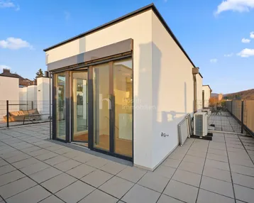 Große Dachterrasse mit Fliesenboden und Zugang zu einem modernen Gebäude unter blauem Himmel.