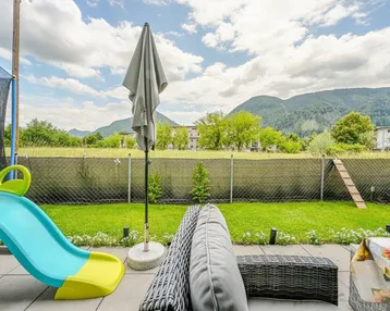 Terrasse mit Kinderrutsche und Bergblick, umgeben von Grünflächen und einem Trampolin.