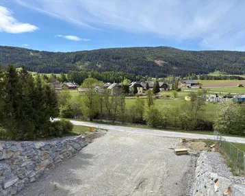 Ländliche Umgebung mit Häusern, grünen Feldern und bewaldeten Bergen im Hintergrund.