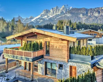 Modernes Chalet mit Holzfassade und Steinelementen vor beeindruckender Bergkulisse an einem sonnigen Tag.
