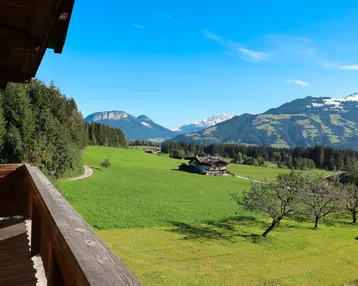 Blick vom Holzbalkon auf eine weite grüne Landschaft mit majestätischen Bergen im Hintergrund unter blauem Himmel.