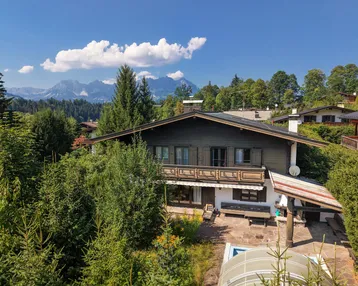 Traditionelles Haus im Alpenstil mit Holzbalkon und Bergblick unter blauem Himmel.