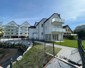 Modernes Neubaugebäude mit hellen Fassaden, Glasbalkonen und angrenzendem Grünbereich am Wasserlauf.
