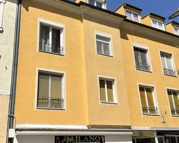 Gelbe Fassade eines Mehrfamilienhauses mit Ladenlokal im Erdgeschoss und mehreren Fenstern.