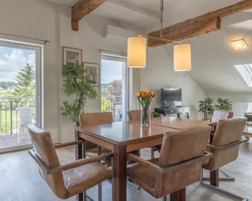 Heller Essbereich mit Holztisch, Ledersesseln, freiliegenden Holzbalken und Zugang zum Balkon mit grüner Aussicht.