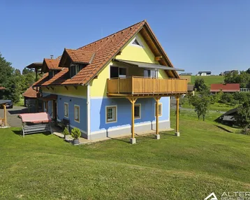 Traditionelles Haus mit gelb-blauer Fassade, Holzbalkon und Satteldach in ländlicher Umgebung.