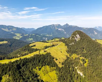 Panoramablick auf eine weite Berglandschaft mit grünen Hügeln, dichten Wäldern und einem klaren blauen Himmel.
