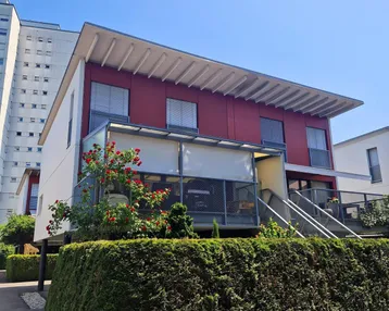 Modernes Gebäude mit markanter roter Fassade, verglaster Loggia und gepflegter Hecke im Vordergrund unter blauem Himmel.