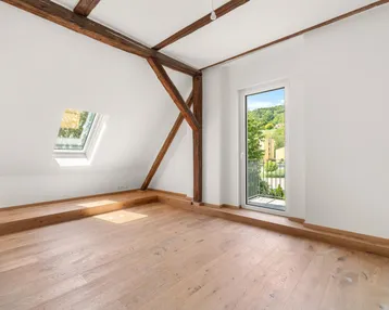 Helles Zimmer mit Holzboden, sichtbaren Holzbalken, Dachfenster und Zugang zu einem französischen Balkon.