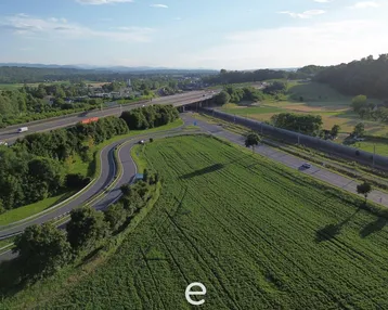 Luftaufnahme einer Autobahnkreuzung, umgeben von grünen Feldern und Wäldern unter blauem Himmel.