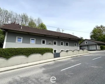Langes Wohnhaus mit grauer Fassade, Hecke und angrenzender Garage an einer Straße.