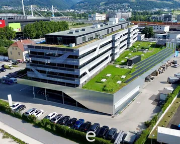 Luftaufnahme eines modernen Bürogebäudes mit begrüntem Dach, Solaranlagen und umliegenden Parkplätzen in städtischer Umgebung.
