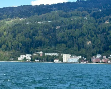 Panoramablick über den See mit Uferbebauung und bewaldeten Hügeln im Hintergrund.