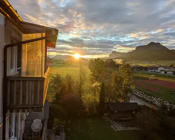 Sonnenuntergang über einer ländlichen Landschaft mit Bergen und einem Balkon im Vordergrund.