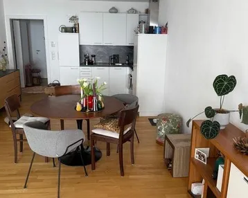 Offener Wohn-Essbereich mit Holzboden und integrierter, moderner Küchenzeile in Weiß.