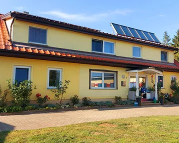 Gelbes Wohnhaus mit rotem Ziegeldach, Solaranlage und gepflegtem Vorgarten unter blauem Himmel.