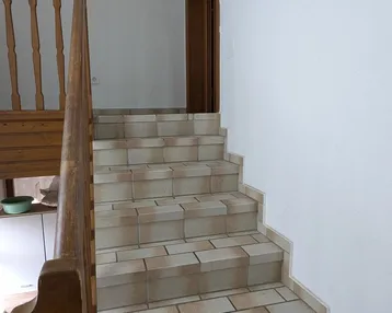 Geflieste Treppe mit Holzgeländer und weißen Wänden, die in den oberen Bereich führt.