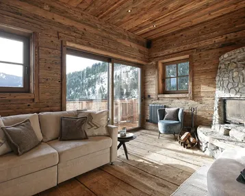 Gemütliches Wohnzimmer im alpinen Stil mit Holzwänden, Steinkamin und Panoramablick auf die verschneiten Berge.