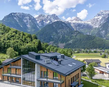 Modernes Gebäude mit Holzfassade und dunklem Dach, eingebettet in eine grüne Landschaft mit majestätischen Bergen im Hintergrund.