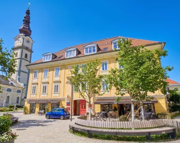 Gelbes Gebäude mit Geschäften im Erdgeschoss und einem Kirchturm im Hintergrund, umgeben von einem belebten Platz.