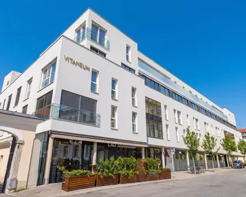 Modernes Mehrzweckgebäude mit weißer Fassade, großen Fenstern und Balkonen in einer städtischen Umgebung.