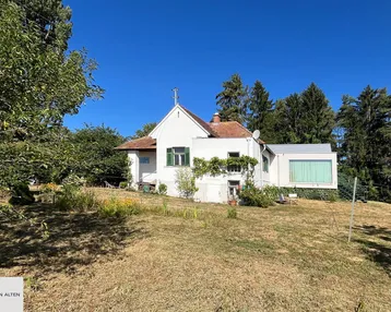 Weißes Haus mit rotem Ziegeldach, grünen Fensterläden und modernem Anbau in ländlicher Umgebung.