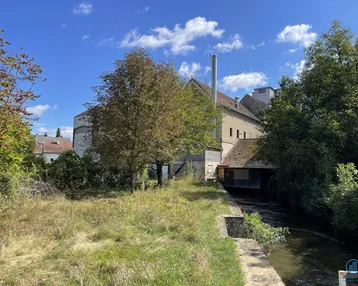Altes Industriegebäude am Flussufer mit viel Grün und einem Schornstein unter blauem Himmel.