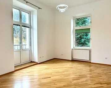 Heller Raum mit Parkettboden, großen Fenstern und Zugang zu einem französischen Balkon.