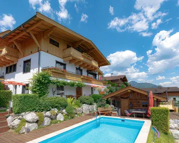 Charmantes Gebäude mit Holzbalkonen, umgeben von einem gepflegten Garten und einem erfrischenden Pool unter blauem Himmel.