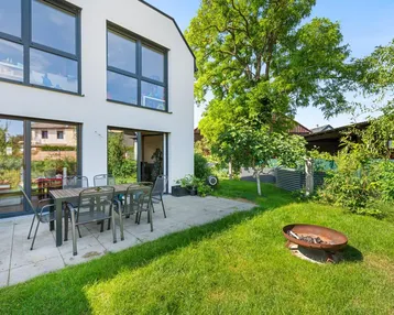 Moderne Hausfassade mit großer gepflasterter Terrasse, Gartenmöbeln und Rasenfläche mit Feuerstelle.