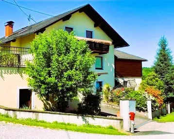 Ansicht des Hauses von außen mit Balkon, umgeben von viel Grün und Bäumen unter blauem Himmel.