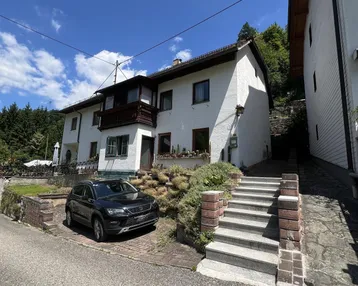 Weißes Wohnhaus mit Balkon und Außentreppe, umgeben von grüner Vegetation unter blauem Himmel.