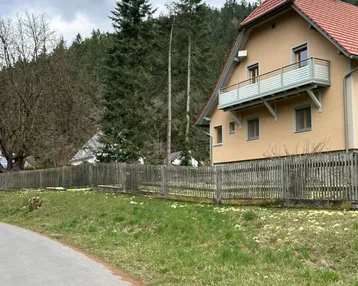 Hausfassade mit Balkon, Holzzaun und bewaldetem Hügel im Hintergrund unter bewölktem Himmel.