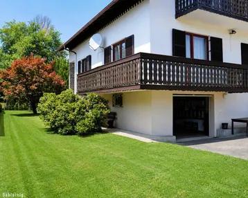 Weißes Haus mit Holzbalkon, gepflegtem Rasen und Bäumen im Gartenbereich.