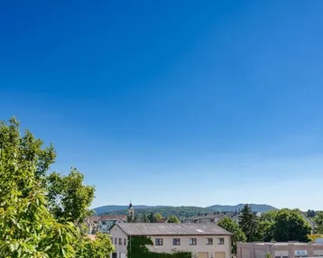 Weitläufiger Blick über die Stadt und die umliegende grüne Landschaft mit Bergen im Hintergrund.