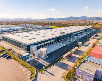 Luftaufnahme eines großen Logistikzentrums mit Laderampen und umliegender Industrielandschaft vor Bergkulisse.