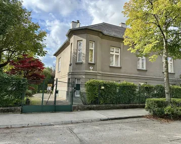 Historisches Gebäude mit klassischer Architektur, umgeben von Hecken und Bäumen an einer ruhigen Straße.