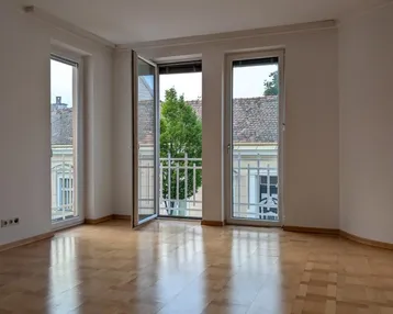 Helles Zimmer mit Parkettboden und Zugang zu einem französischen Balkon mit Blick auf die Straße.