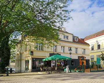 Gebäude mit Außengastronomie und Kinderspielplatz an einem sonnigen Tag, umgeben von Bäumen und einem Platz.