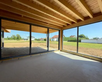 Unfertiger Innenraum mit Holzbalkendecke und großen Schiebetüren zu einem zukünftigen Gartenbereich.