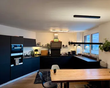 Moderne Küche mit schwarzen Fronten, integrierten Geräten und Holztisch, beleuchtet von stilvollen Hängelampen.